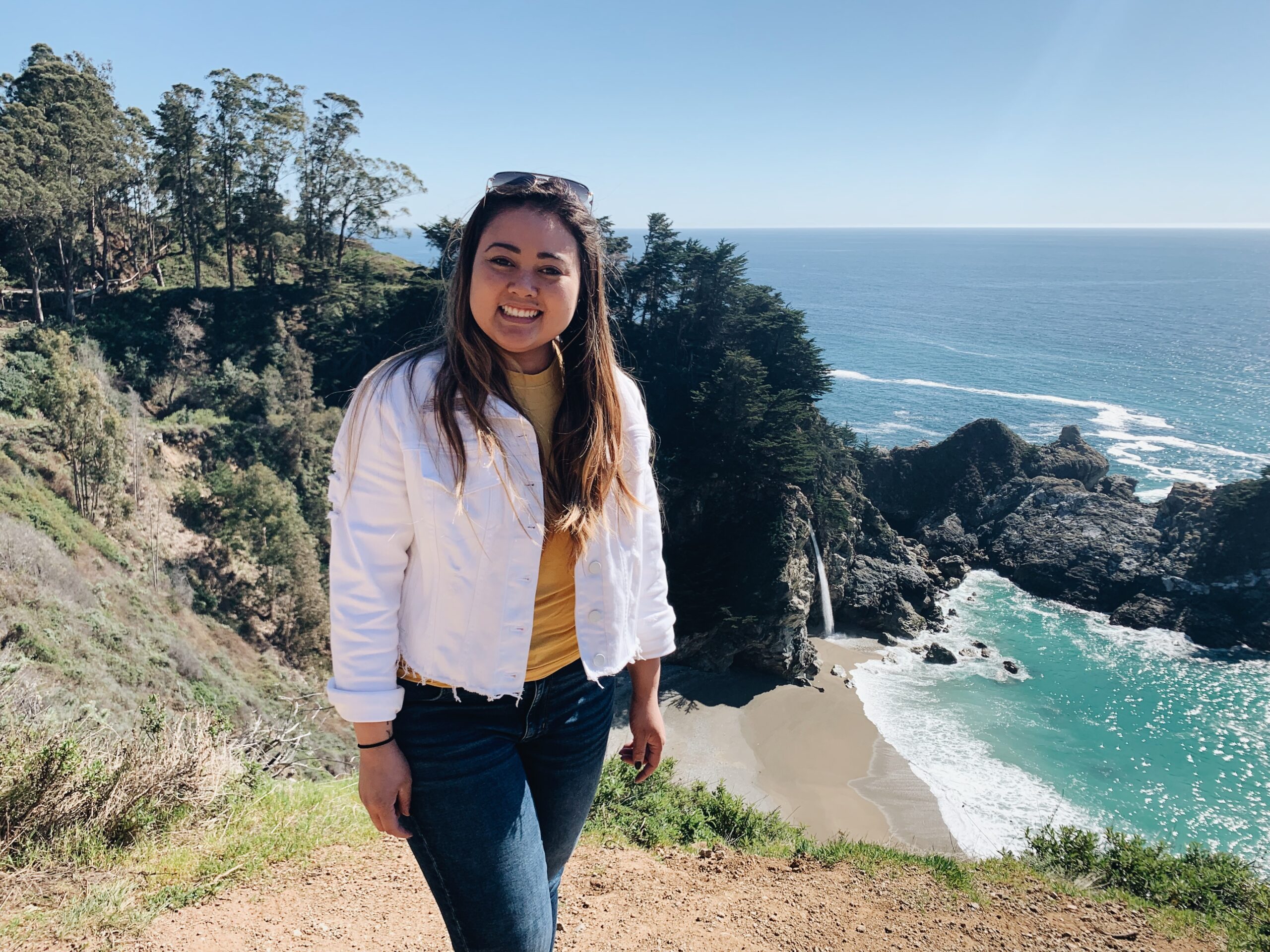 Mcway falls, California girl smiling and posing in front of mcway falls in california wearing a white denim jacket, yellow top, and blue jeans.