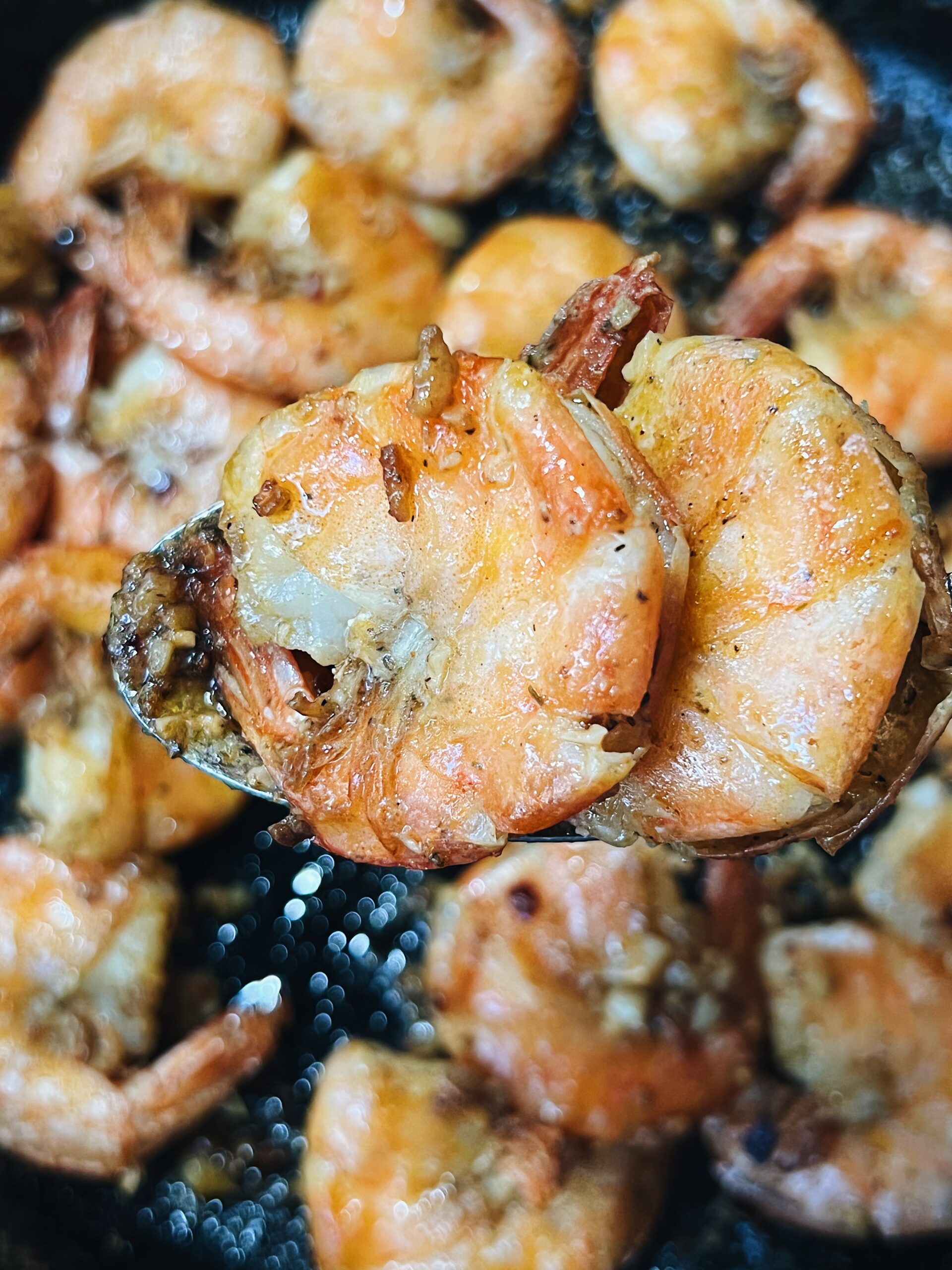 2 garlic shrimp cooked with shell on in a spoon being held up to the camera. garlic shrimp in a pan in the background.