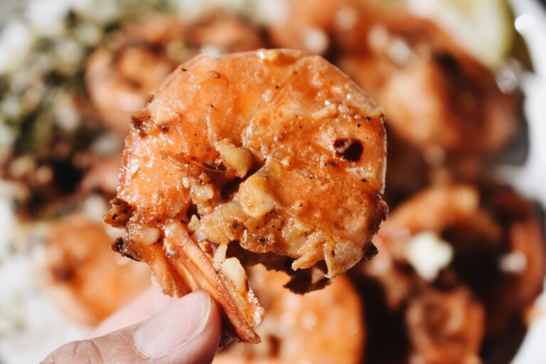 cooked garlic shrimp with shell on held up to the camera with a hand and other shrimp in the background