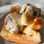 a big hollow piece of toast sitting on a plate with cubes of baked butter honey toast inside topped with 2 scoops of cookies and cream ice cream and honey drizzled on top.