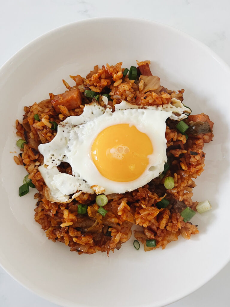 Riced mixed with Kimchi, eggs, spam, garlic, onion, and green onion with sauce to make Kimchi Fried rice. Kimchi Fried Rice in a bowl with a sunny side up egg on top.
